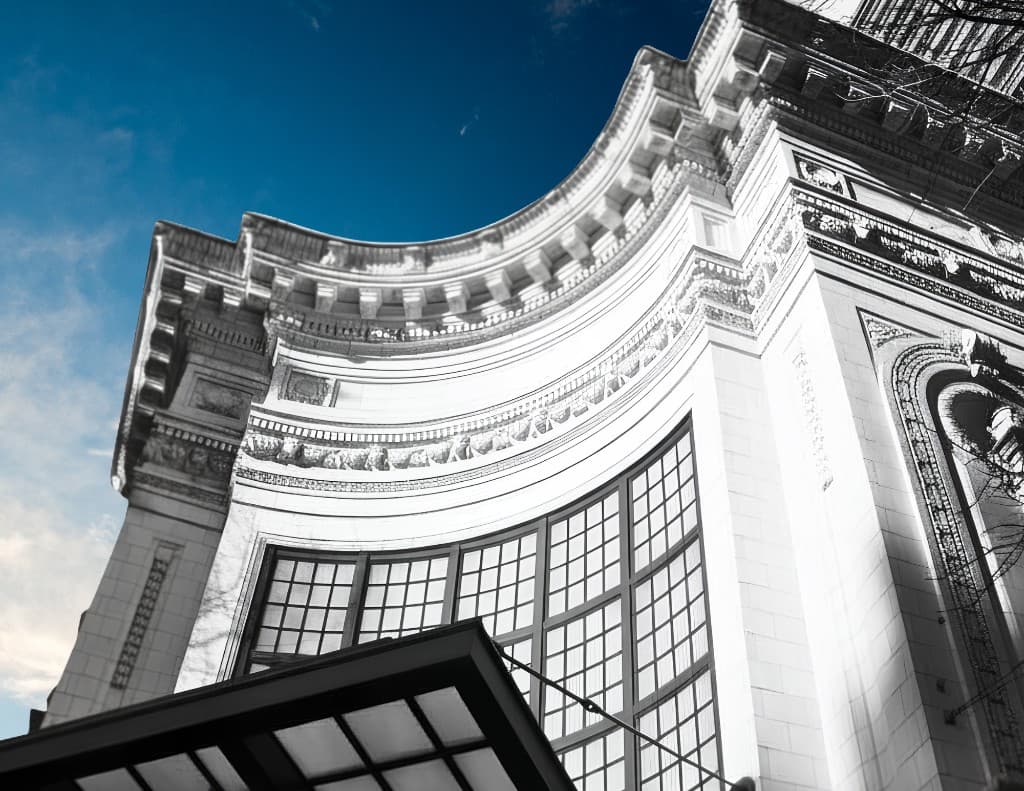 Low-angle view of the curved Coliseum exterior with ornate stonework, tall grid windows, and blue sky overhead