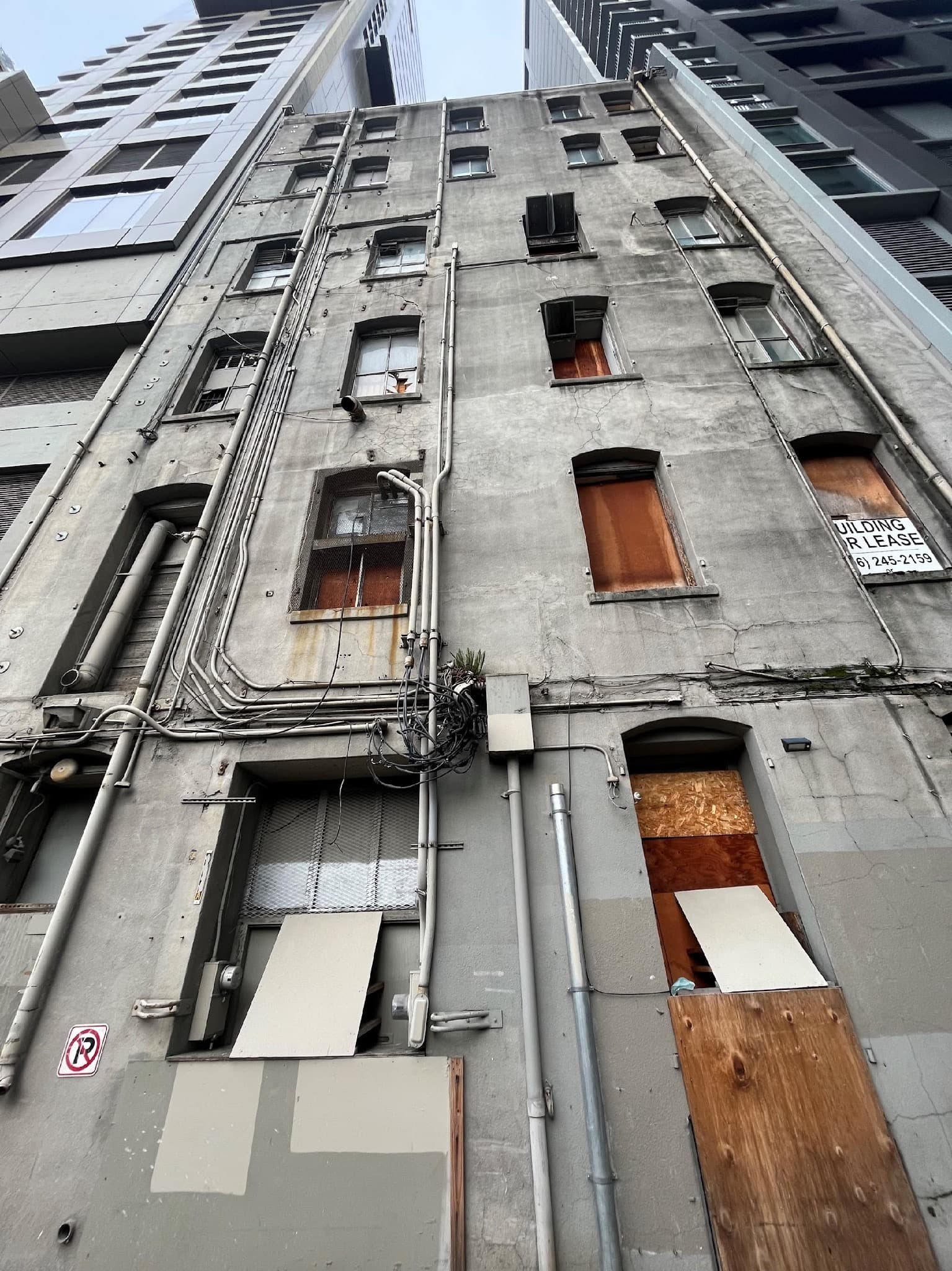 The rear facade of the Seven Seas Building, a tall weathered concrete-and-brick back wall with boarded windows and a faded Building For Lease sign, dropping toward Post Alley