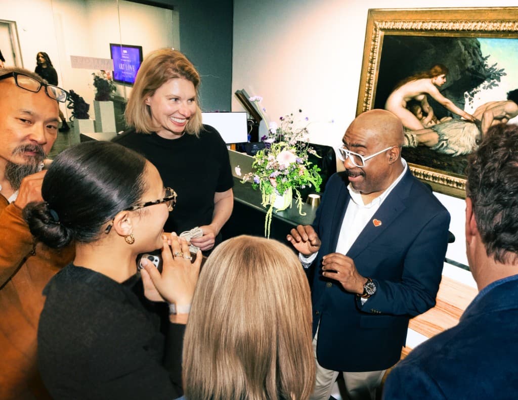 A small group of guests talk and smile near a desk area, with a large framed painting in the background.