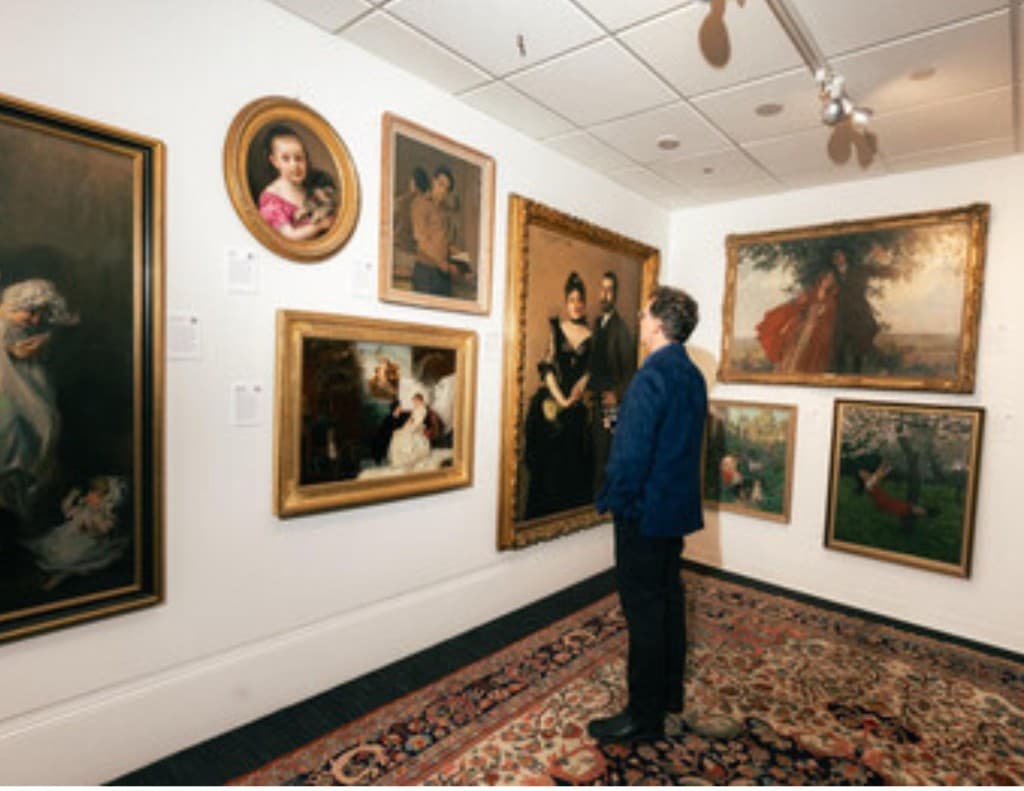 A visitor in a blue jacket stands on a patterned rug and looks at framed portrait paintings in a white gallery corner.