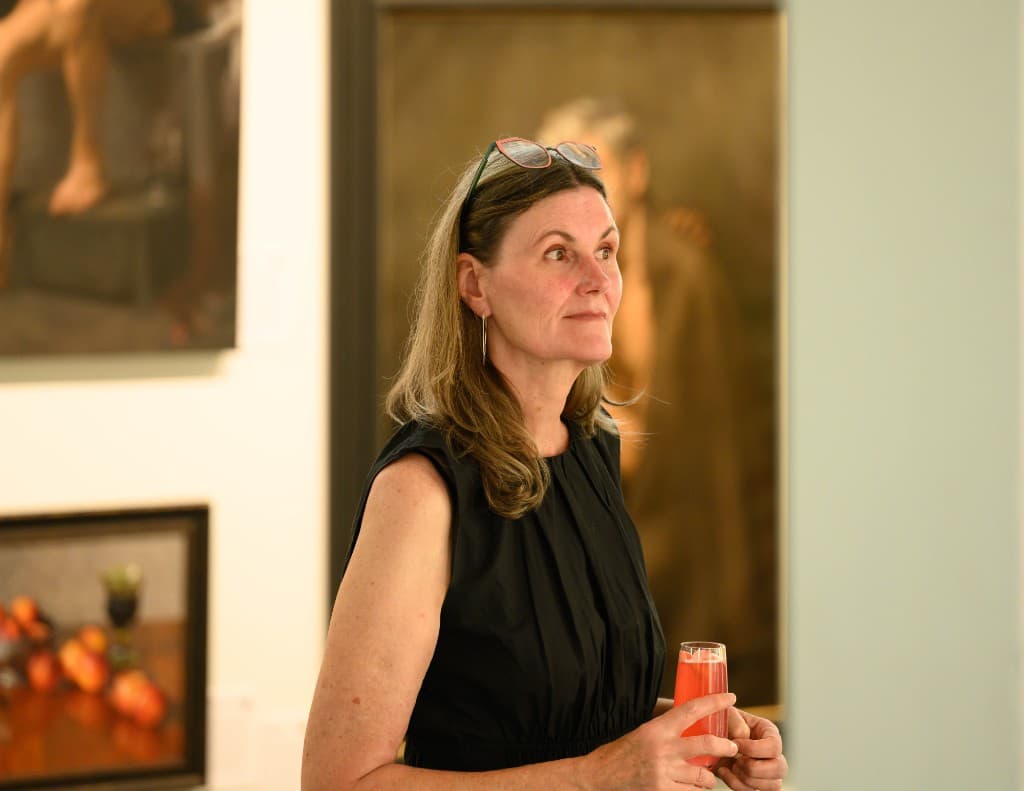 A guest in a black sleeveless dress holds a pink drink while standing among framed paintings at an evening reception.