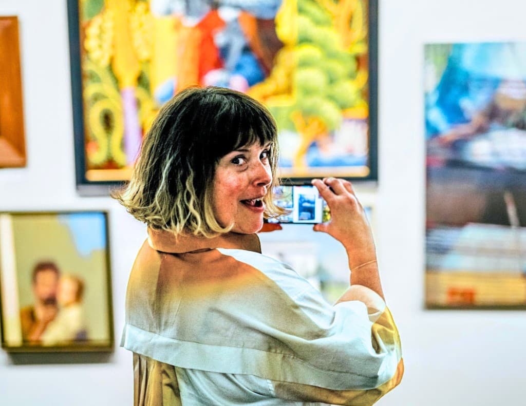 A smiling guest turns toward the camera while holding a phone for a selfie, with colorful paintings behind her.