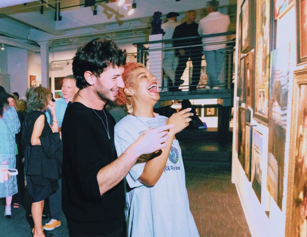 Two guests laugh together while looking at a wall of framed artwork during a crowded evening salon event.
