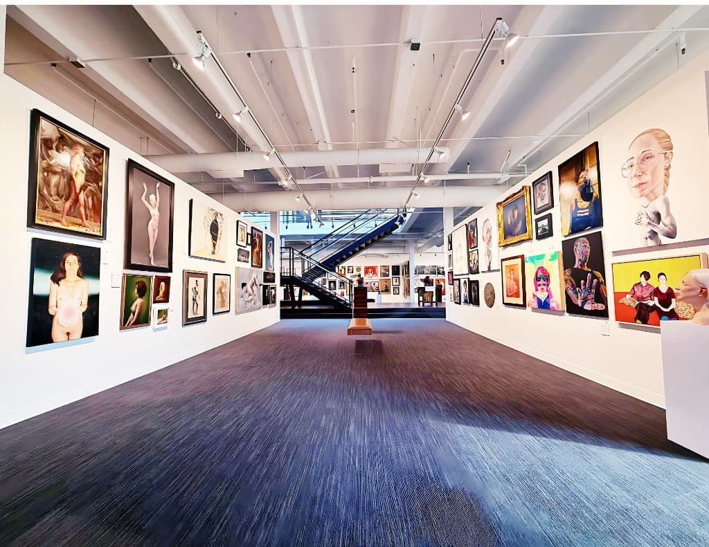A wide-angle view of a bright gallery floor with many paintings on both walls, open stairs in the center, and overhead lighting.