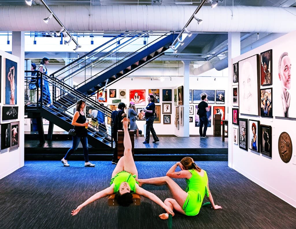 Two performers in bright green outfits pose on the gallery floor while visitors and artwork fill the multi-level space behind them.
